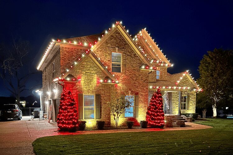 A large, upscale brick home beautifully decorated with festive outdoor Christmas lighting at night.