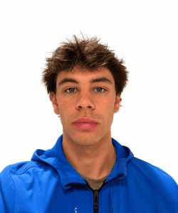 A young man with tousled brown hair wearing a bright blue zip-up jacket looks directly at the camera against a white background.
