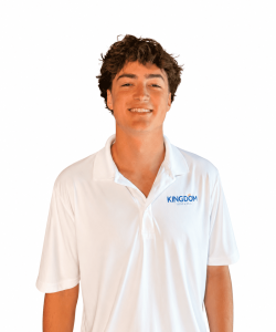 Young man with brown hair wearing a white polo shirt that has "KINGDOM" written on it, standing and smiling at the camera.