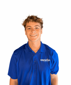 Young man in a blue "KINGDOM" polo shirt stands facing forward and smiling against a plain white background.