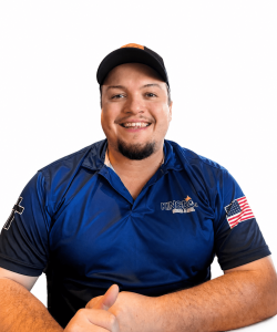Smiling man in a blue "Kingdom" polo shirt and black cap sitting at a table, with a cross and USA flag on his sleeves.