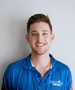 A man in a blue "Kingdom" polo shirt stands against a plain white wall, smiling at the camera.