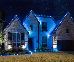 A two-story house decorated with blue and white lights along the roof, windows, and landscaping at night.