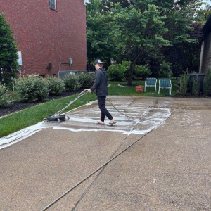 Person pressure washing a soapy driveway with a surface cleaner in a residential outdoor area.