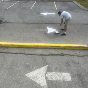 Person painting a white arrow on a parking lot surface near yellow curb; other arrows point different directions.