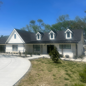 Single-story white house with black roof, three dormer windows, front porch, and a curved driveway under a clear sky.