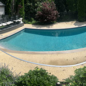 A clear swimming pool surrounded by a concrete deck, landscaping, and seating area with chairs in the background.