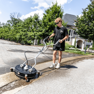 A person uses a large surface cleaner to wash a paved street near a residential area on a sunny day.