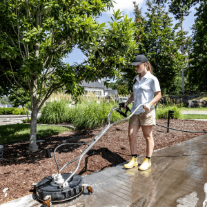 A person uses a pressure washer to clean a concrete walkway in a landscaped outdoor area.