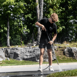A person wearing black clothes uses a pressure washer to clean an outdoor pavement with trees and rocks in the background.