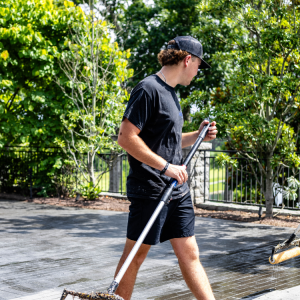 Person in black shirt and shorts using a long-handled tool to clean an outdoor surface near trees and a metal fence.