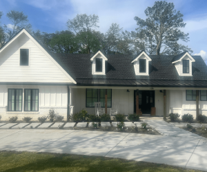 A white modern farmhouse with black trim, dormer windows, and two rocking chairs on the front porch under a blue sky.