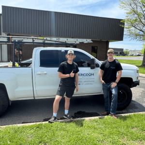 Two men in black shirts stand by a white work truck with a ladder and company logo in a parking lot.