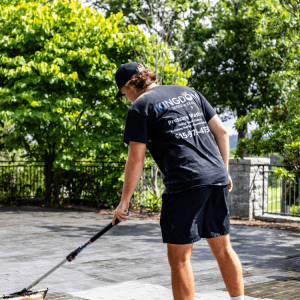 Person wearing a black shirt and shorts uses a long-handled tool to clean an outdoor paved area on a sunny day.