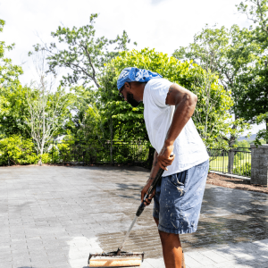 Man wearing a blue bandana and white shirt uses a roller brush to seal a paved outdoor surface on a sunny day.