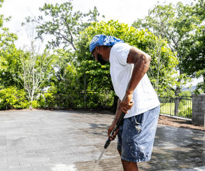 Man wearing a blue headscarf and white shirt uses a roller to apply coating on a paved outdoor surface.