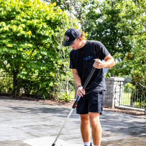 A person wearing a cap and shorts uses a broom to clean an outdoor paved area on a sunny day.