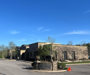 A brick building labeled "Genesis Campus for Jewish Life" with trees, a parking lot, and two orange cones in front.