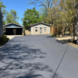 Freshly paved driveway leading to two garages, surrounded by trees and fences under a clear blue sky.